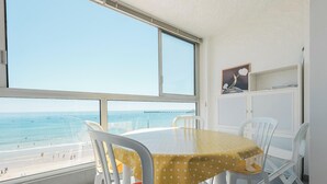A room with a large window overlooking a beach, a table with chairs, and a white cabinet with a picture frame. - Renovated 66m² apartment, 1 bedroom, sea view, 4th floor, fiber, 3 stars (Les Sables-d'Olonne)