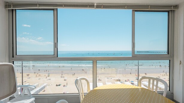 A room with a large window overlooking a beach. There are white chairs and a table with a yellow polka dot tablecloth. - Renovated 66m² apartment, 1 bedroom, sea view, 4th floor, fiber, 3 stars (Les Sables-d'Olonne)