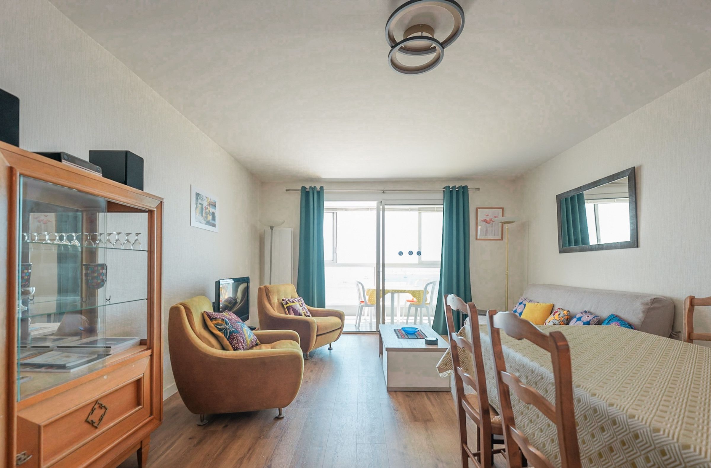 A spacious room with a wooden cabinet, two armchairs, a sofa, a coffee table, and a large window with curtains.