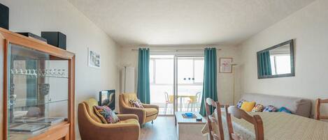A spacious room with a wooden cabinet, two armchairs, a sofa, a coffee table, and a large window with curtains.