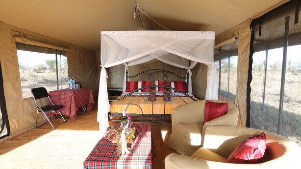 A tented room with a bed, a table with a red and black checkered tablecloth, and a sofa with red cushions. - Serengeti Wild Camps (Serengeti)