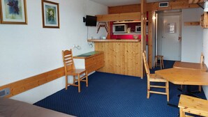 A compact room with a wooden desk, a chair, a dining table with chairs, a kitchenette with a microwave, and a small refrigerator.