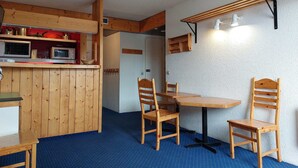 A compact kitchen with wooden cabinets, a microwave, and a hanging rack. There is a dining area with a table and two chairs.