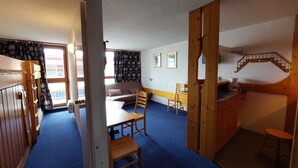 A room with a bunk bed, a sofa, a desk, and a chair. There is a window with curtains, a staircase, and a wall-mounted shelf.