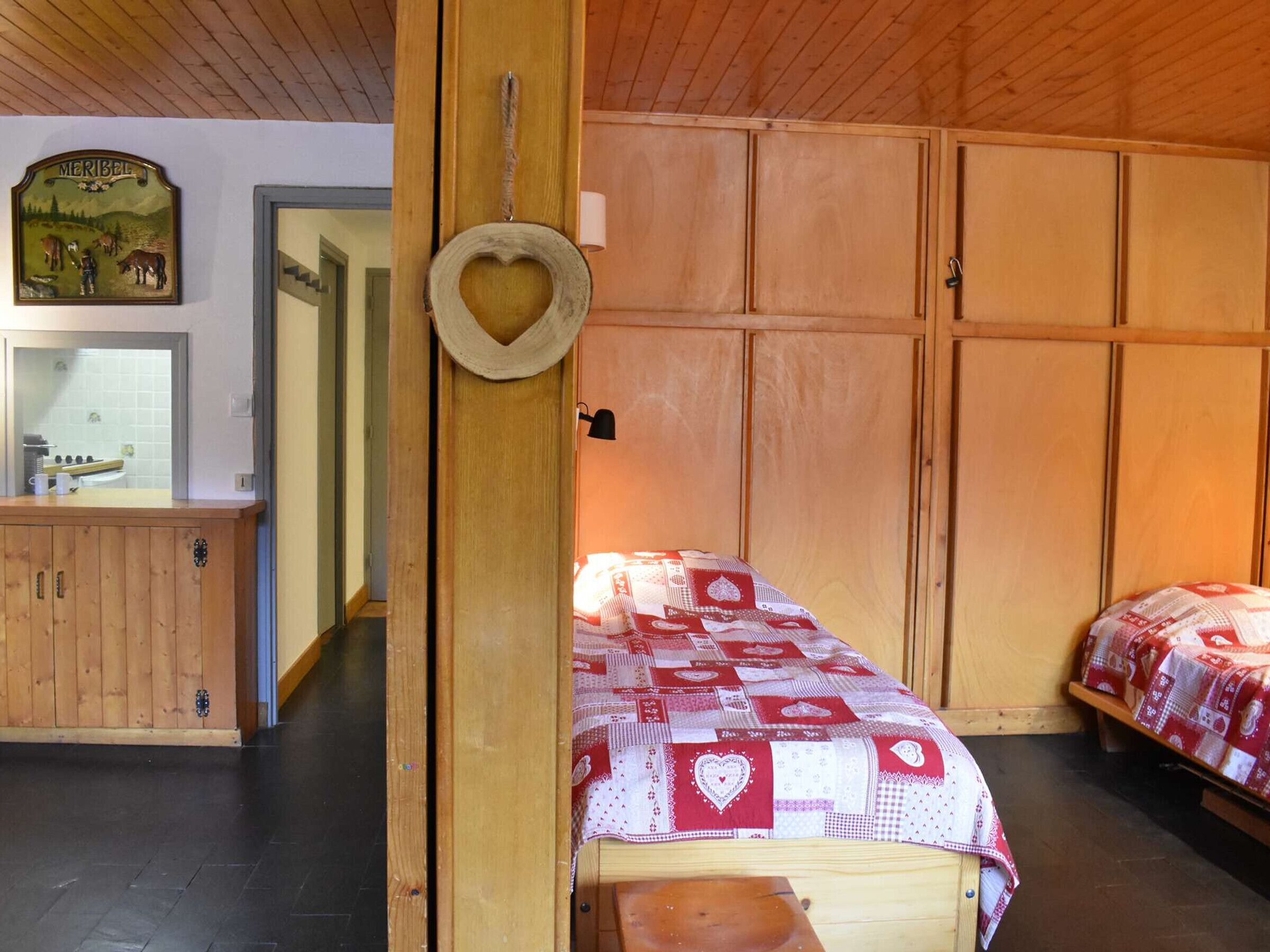 A cozy room with wooden walls and a bed with a red and white quilt. There is a wooden cabinet with a mirror and a picture of a farm scene.
