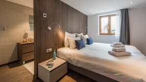 Un dormitorio moderno con una pared de acento de madera, una cama grande con ropa de cama blanca y azul, una mesita de noche con lámpara, y una ventana con cortinas.