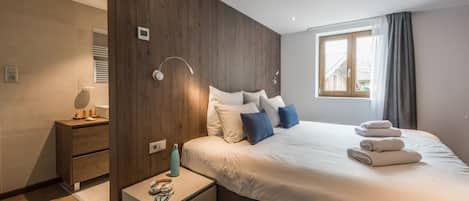 Quarto moderno com uma parede de destaque em madeira, uma cama grande com roupa de cama branca e azul, uma mesa de cabeceira com abajur e uma janela com cortinas.