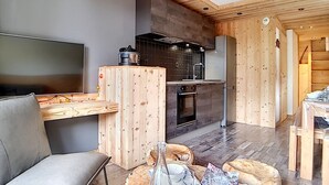 A modern living room with a sofa, a coffee table, and a TV. There is a kitchen area with a stove, oven, and sink. The room has wooden walls and ceiling.