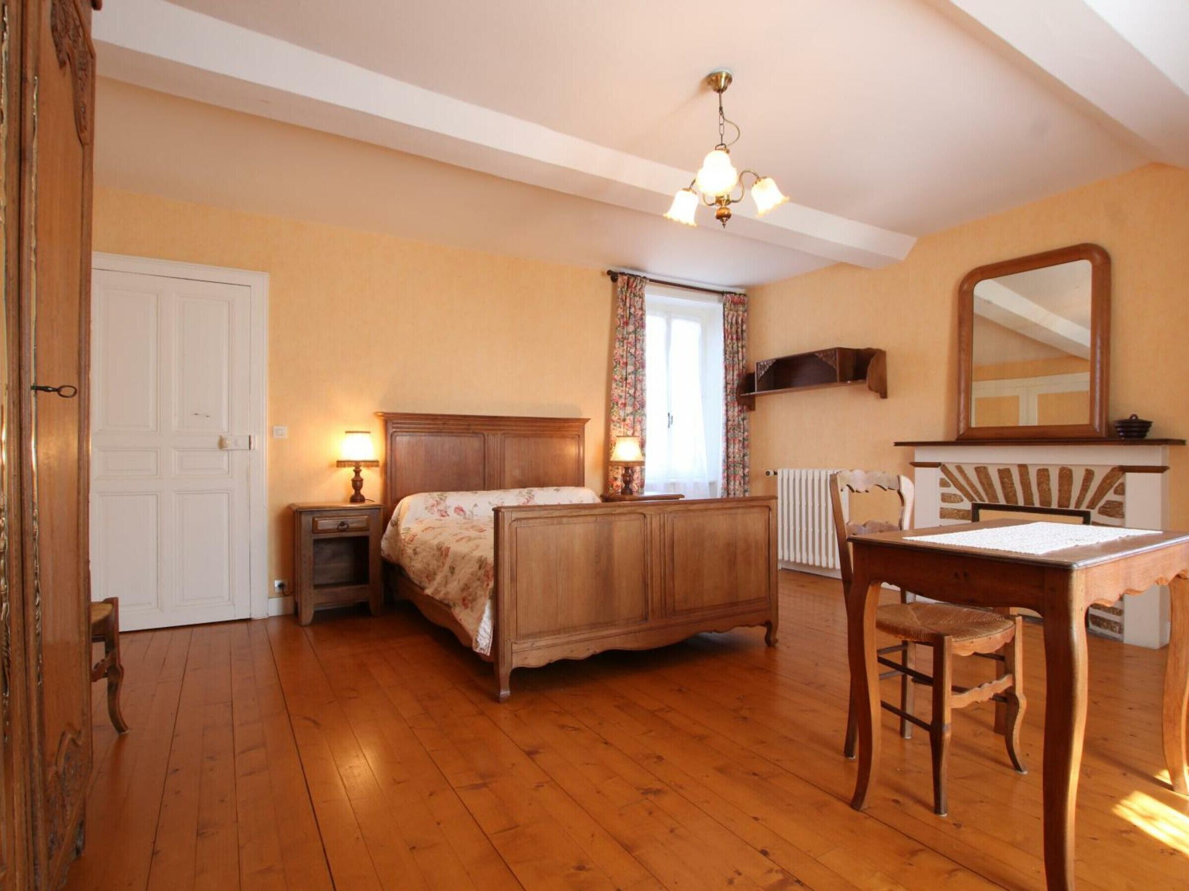 Ein Schlafzimmer mit einem hölzernen Bett, einem Schreibtisch und einem Stuhl. Es gibt ein Fenster mit Vorhängen, eine Heizung und einen Spiegel an der Wand.