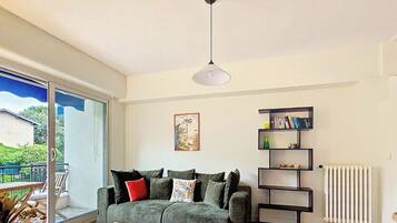 Een woonkamer met een bank, een boekenkast, een radiator en een raam met uitzicht op groen.