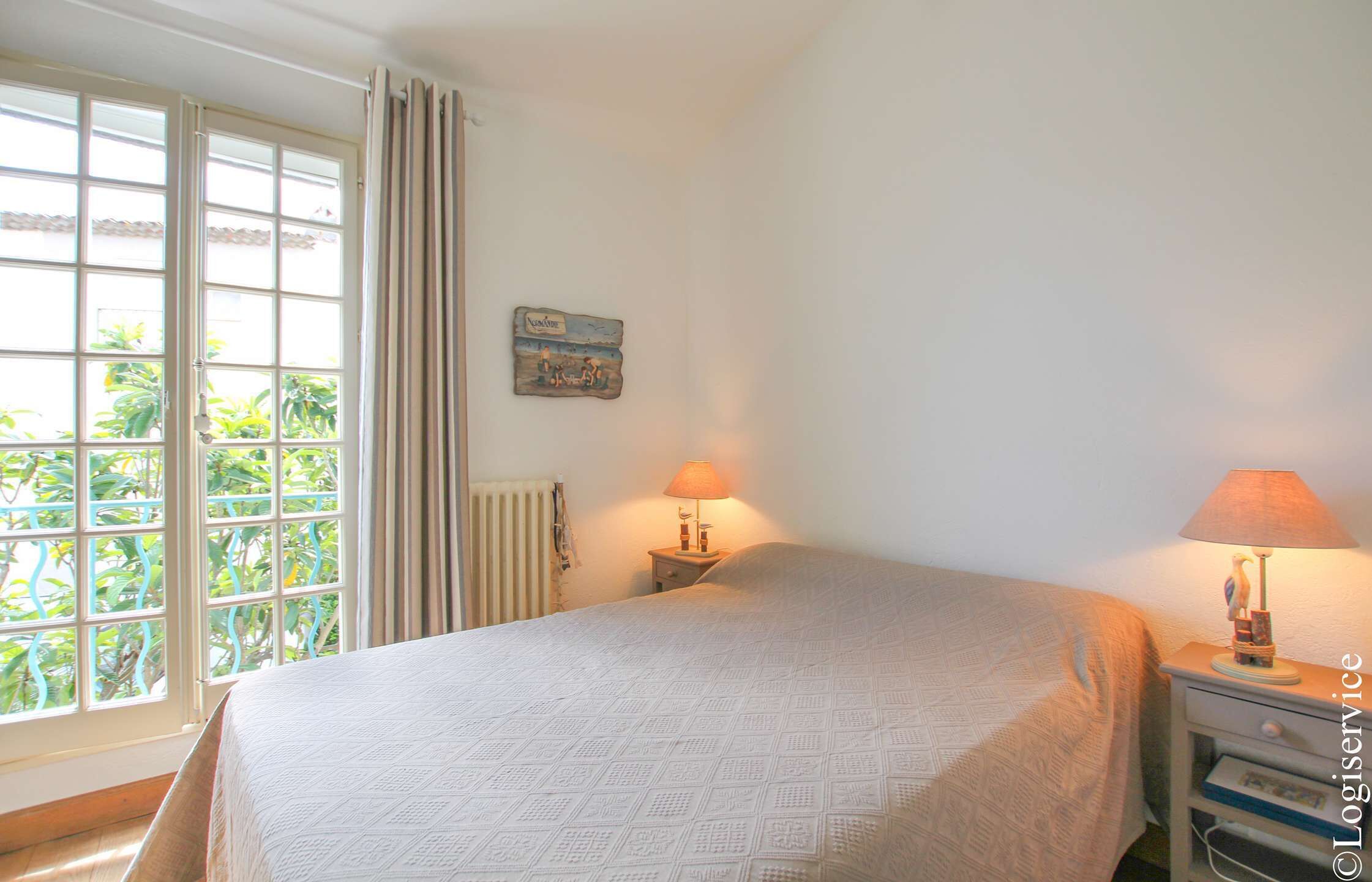 A bedroom with a bed, two bedside tables with lamps, a window with curtains, a radiator, and a small wall decoration.
