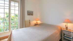 Un dormitorio con una cama, dos mesitas de noche con lámparas, una ventana con cortinas, un radiador y un pequeño adorno en la pared.
