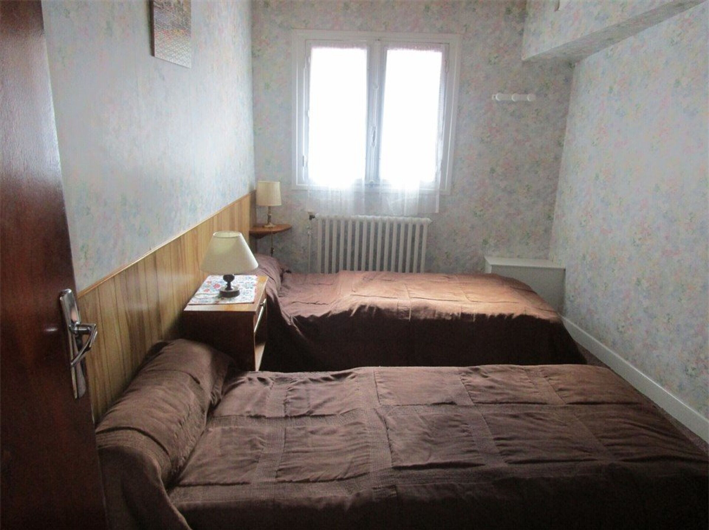 A small, dimly lit room with a single bed, a nightstand with a lamp, and a window with blinds.