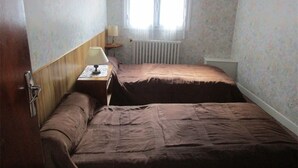 A small, dimly lit room with a single bed, a nightstand with a lamp, and a window with blinds.