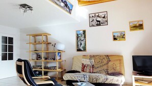 A living room with a wooden ceiling, a sofa with a map-patterned cover, a TV stand, a bookshelf, and a window. - Beautiful 67m² apartment, 2 bedrooms, parking, south-facing balcony - Briançon (Briançon)