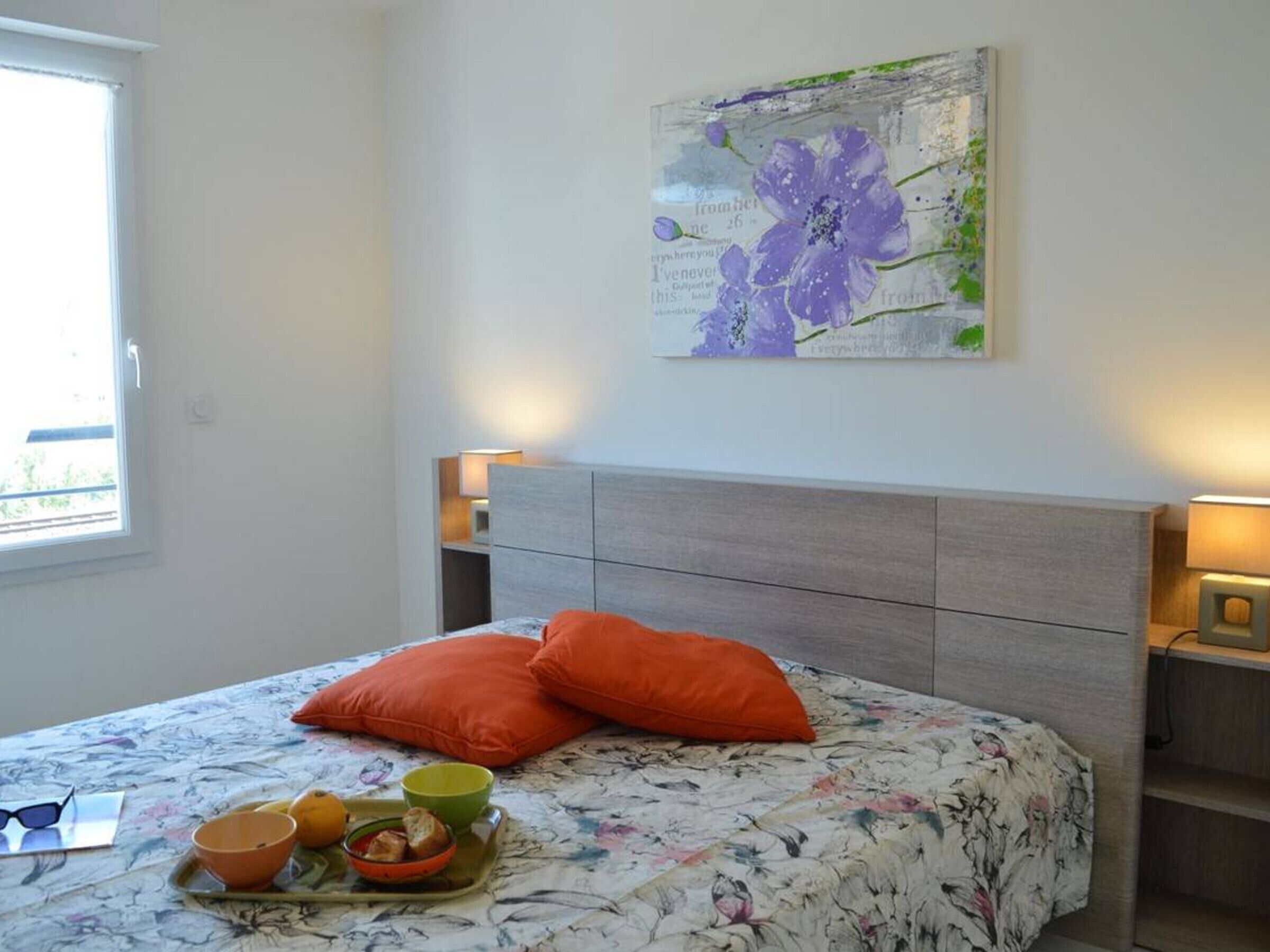 Ein Schlafzimmer mit einem Bett, einem blumigen Bettüberwurf, zwei orangenen Kissen, einer Ablage mit Schüsseln und Brot sowie einem Bild an der Wand.