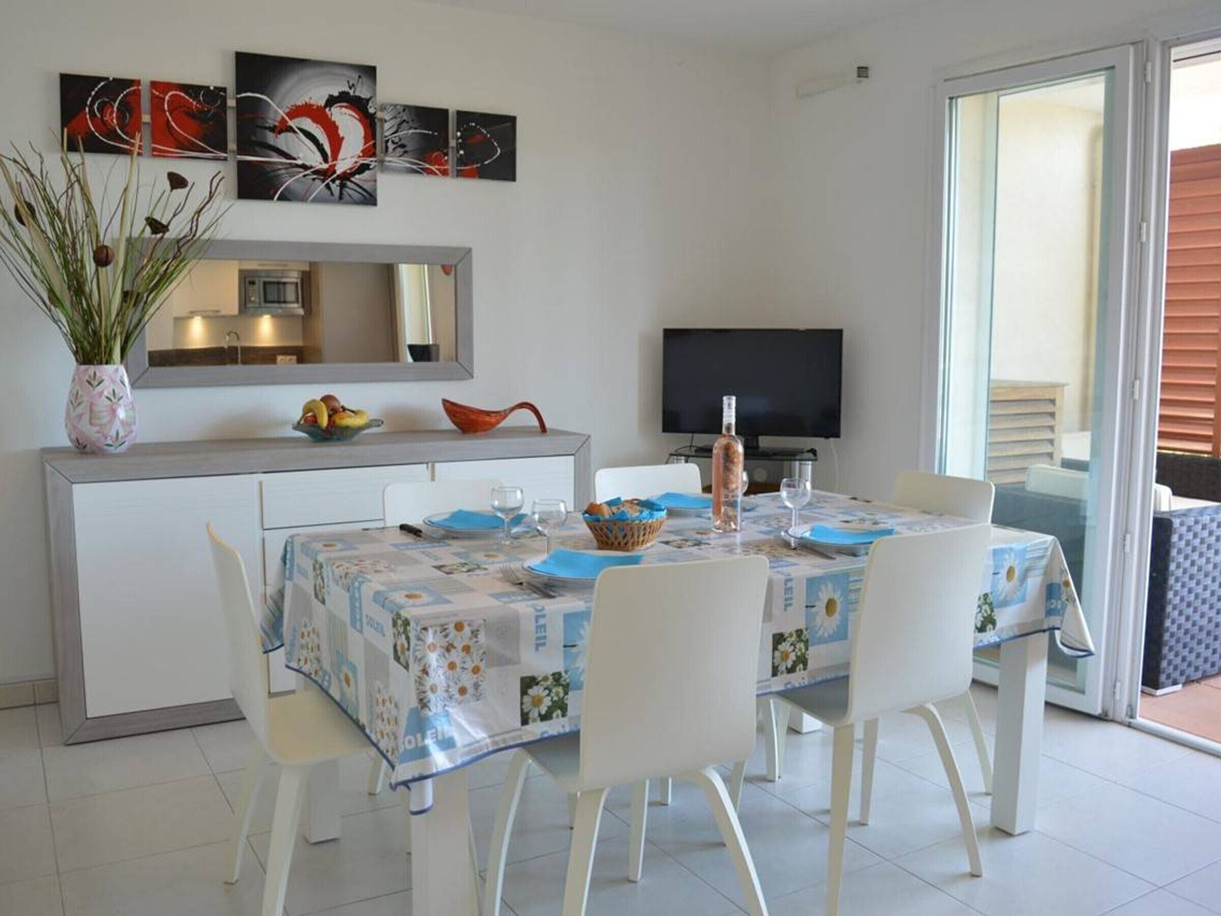 Un coin repas avec une table dressée pour quatre personnes, une plante en pot, un panier de fruits et un téléviseur en arrière-plan.