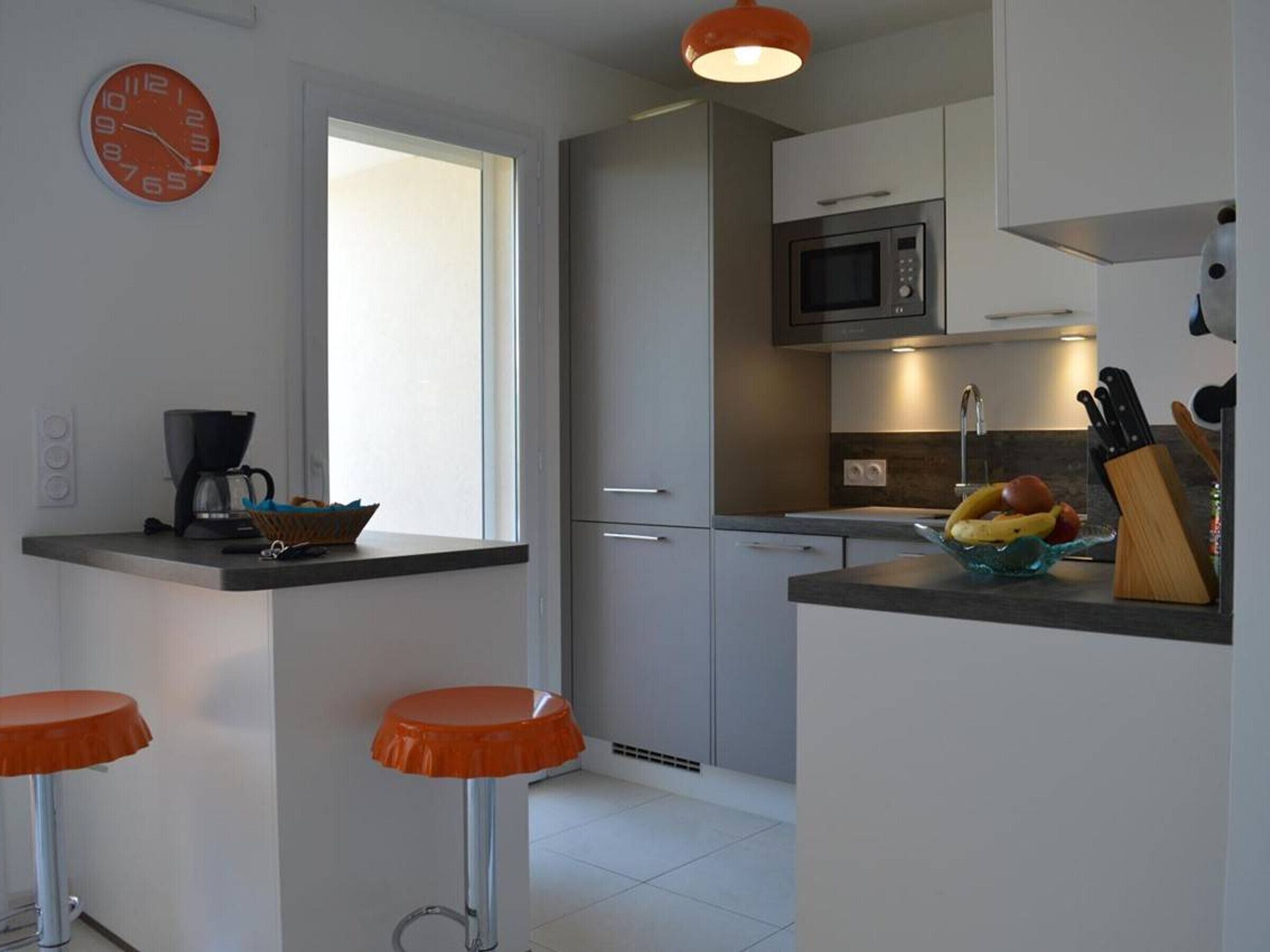 Une cuisine moderne équipée d’un four à micro-ondes, d’une machine à café et d’un panier à fruits posé sur le comptoir. On y trouve également deux tabourets orange et une fenêtre qui laisse entrer la lumière naturelle.