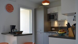 Eine moderne Küche mit Mikrowelle, Kaffeemaschine und Obstschale auf der Arbeitsfläche. Zwei orangefarbene Hocker und ein Fenster sorgen für natürliches Licht.