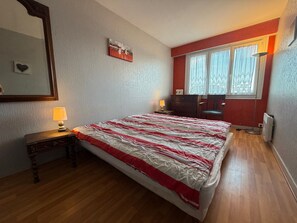 Une chambre à coucher avec un lit, des tables de chevet, une commode, un miroir et une fenêtre avec des rideaux.