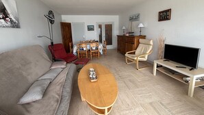Una sala de estar con un sofá, una mesa de centro de madera, un mueble para la televisión, un sillón rojo, una mesa de comedor con sillas y un armario.
