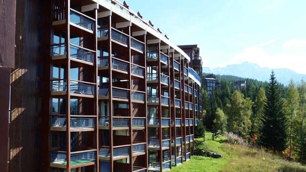 A multi-story hotel with balconies, surrounded by greenery and mountains.