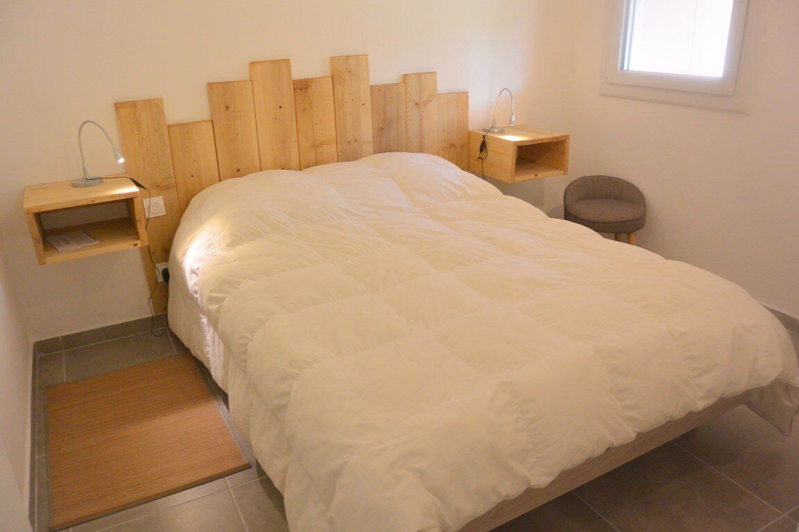 Ein Schlafzimmer mit hölzernem Kopfteil, einem Bett mit weißer Bettwäsche, einem Nachttisch mit Lampe, einem Stuhl und einem kleinen Regal.
