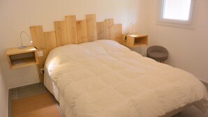 A bedroom with a wooden headboard, a bed with white bedding, a bedside table with a lamp, a chair, and a small shelf. - MINI-VILLA SAINT CYPRIEN C20- 3***-QUIET (Saint-Cyprien Lecci)