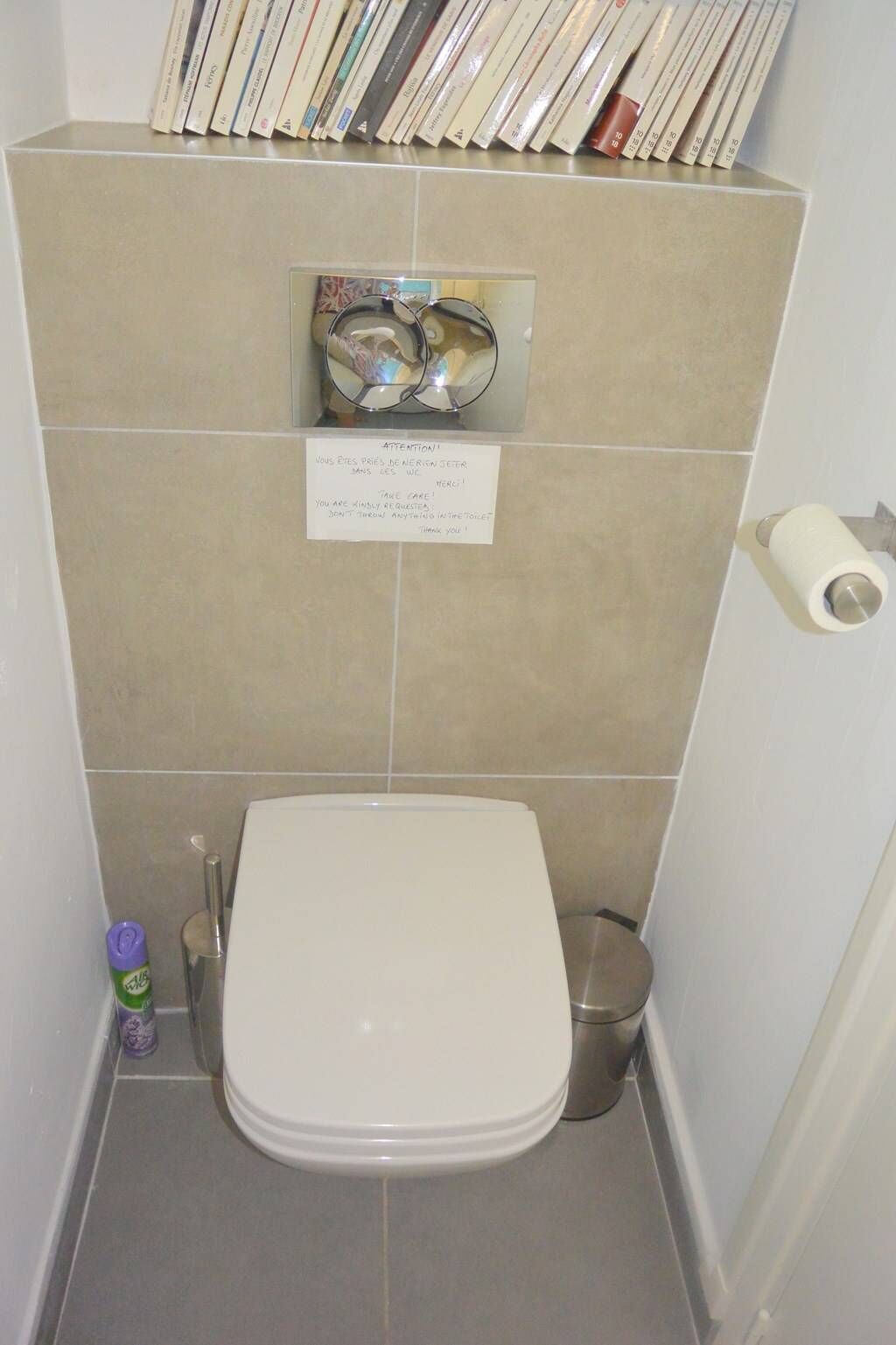 A small bathroom with a toilet, a trash bin, and a toilet paper holder. There is a sign with text in multiple languages.