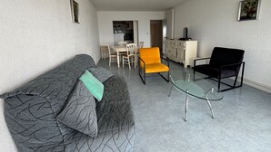 A living room with a grey patterned sofa, an orange armchair, a black armchair, and a glass coffee table. There is a dining area with a white table and chairs, and a kitchen in the background. - 2-Bedroom Apartment with Balcony and Garage in Les Sables-d'Olonne (Les Sables-d'Olonne)