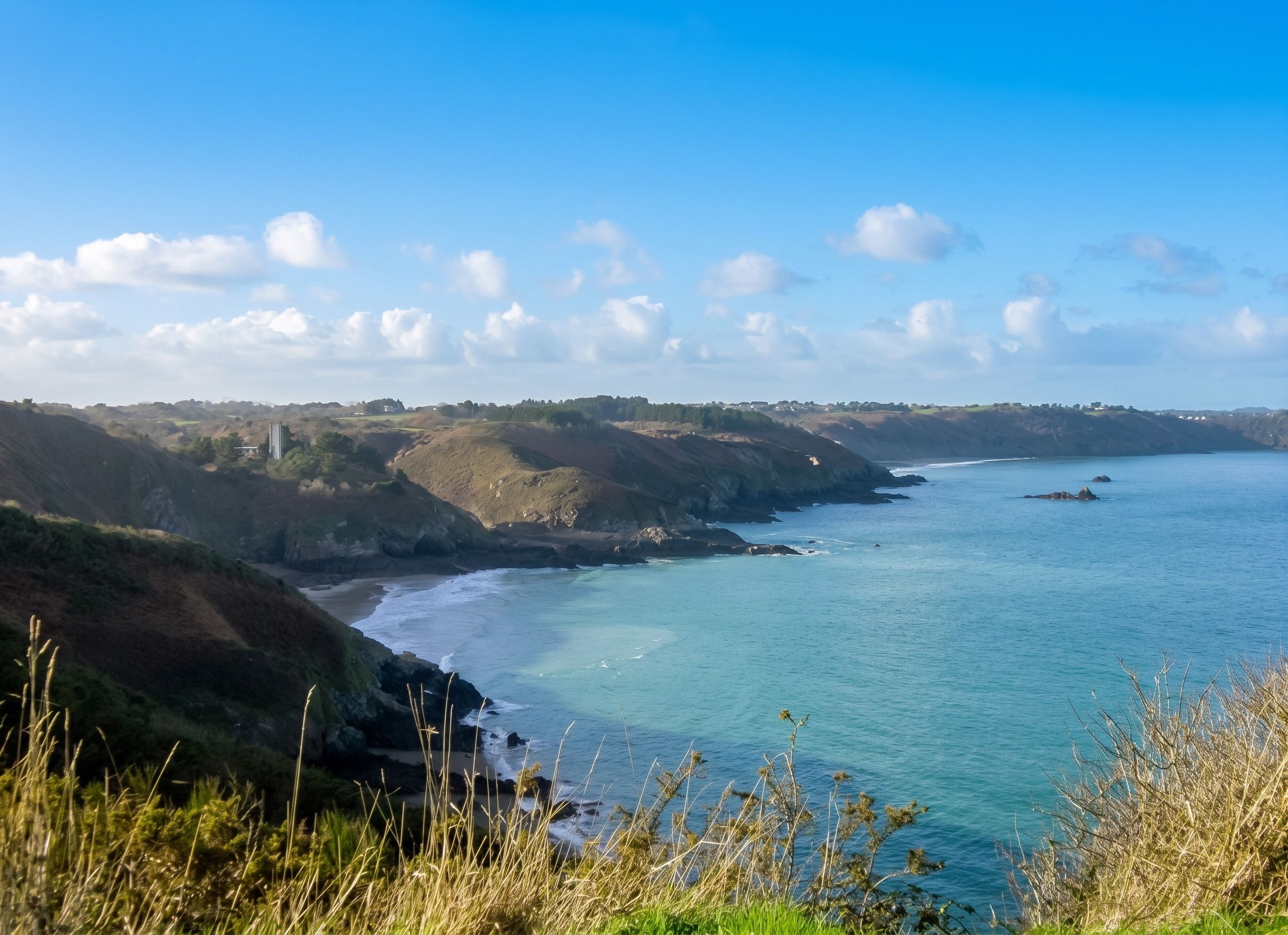 A coastal landscape with a clear sky, a calm sea, and rolling hills.