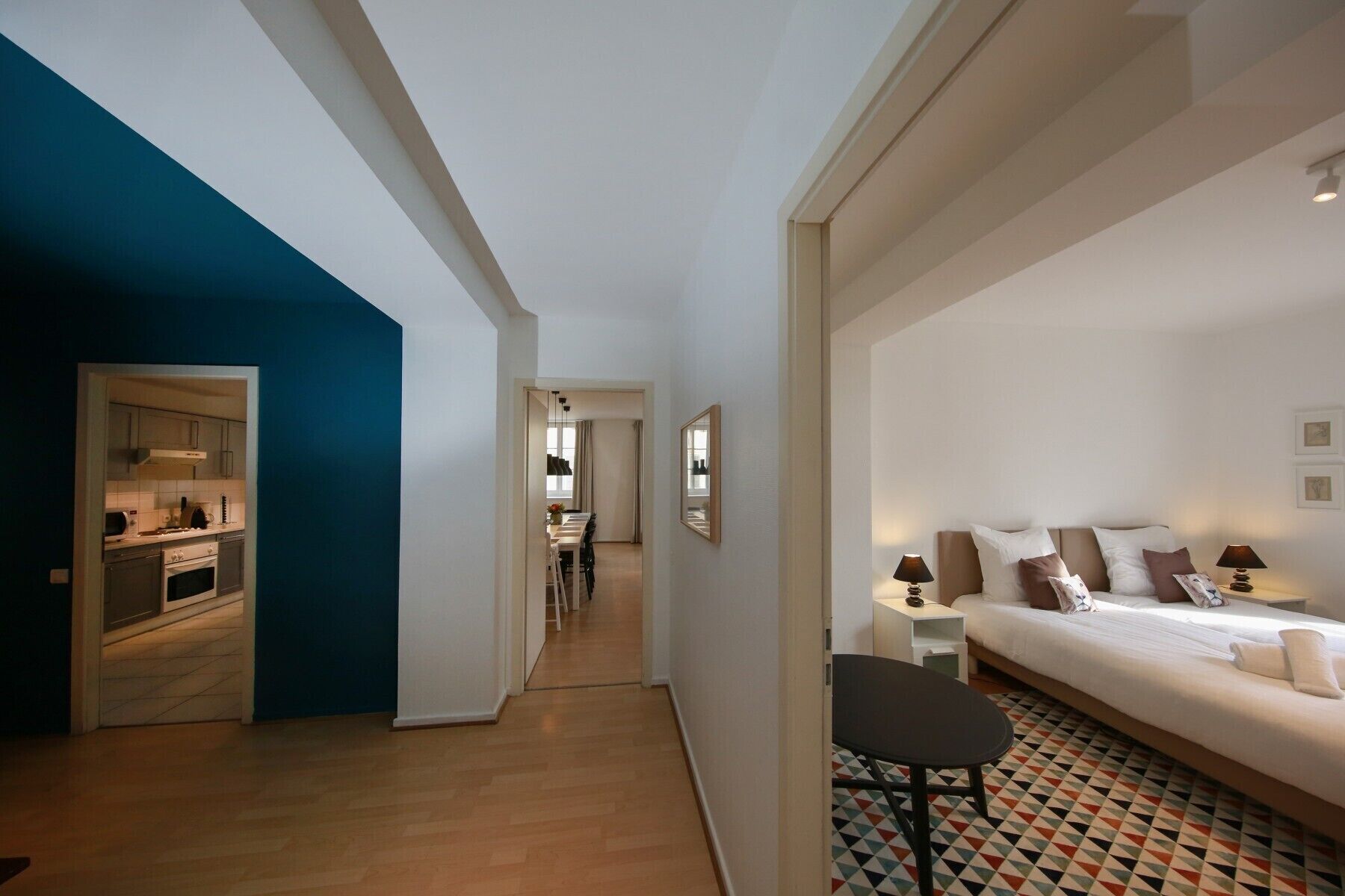 A modern hotel room with a large bed, a small table, and a chair. There is a kitchen area visible through an open door.