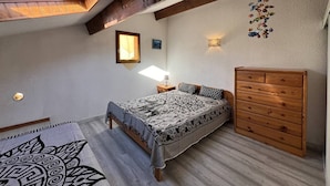 Un dormitorio con techo inclinado, una cama con ropa de cama estampada, un aparador de madera y una lámpara de pared.