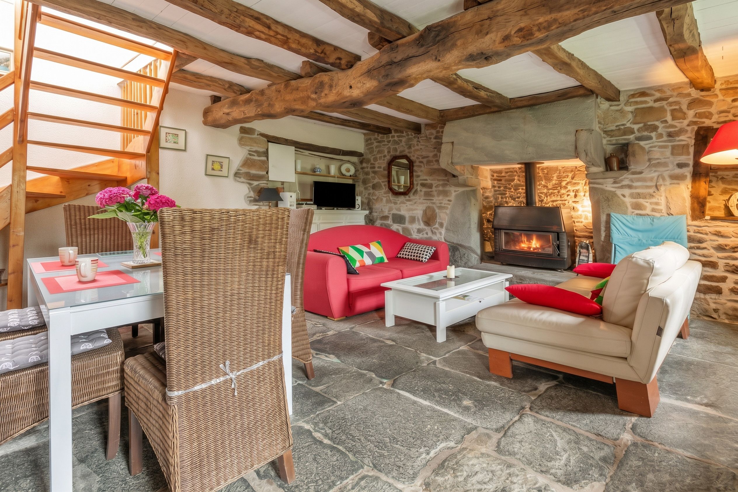 Ein gemütliches Wohnzimmer mit Kamin aus Stein, einem roten Sofa, einem Korbstuhl und einem Essbereich mit Glastisch.