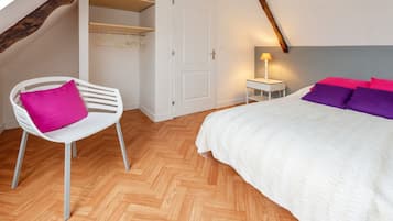 A bedroom with a herringbone wooden floor, a white bed with purple and pink pillows, a white chair with a pink cushion, and a bedside table with a lamp.