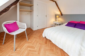 A bedroom with a herringbone wooden floor, a white bed with purple and pink pillows, a white chair with a pink cushion, and a bedside table with a lamp. - Homerez - Charming House in Guimaëc, Sea View (Guimaëc)