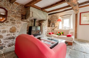 Ein Zimmer mit Steinwänden, einem Kamin, einem roten Sessel, einem weißen Sofa mit bunten Kissen und einem Fenster mit einer roten Lampe.