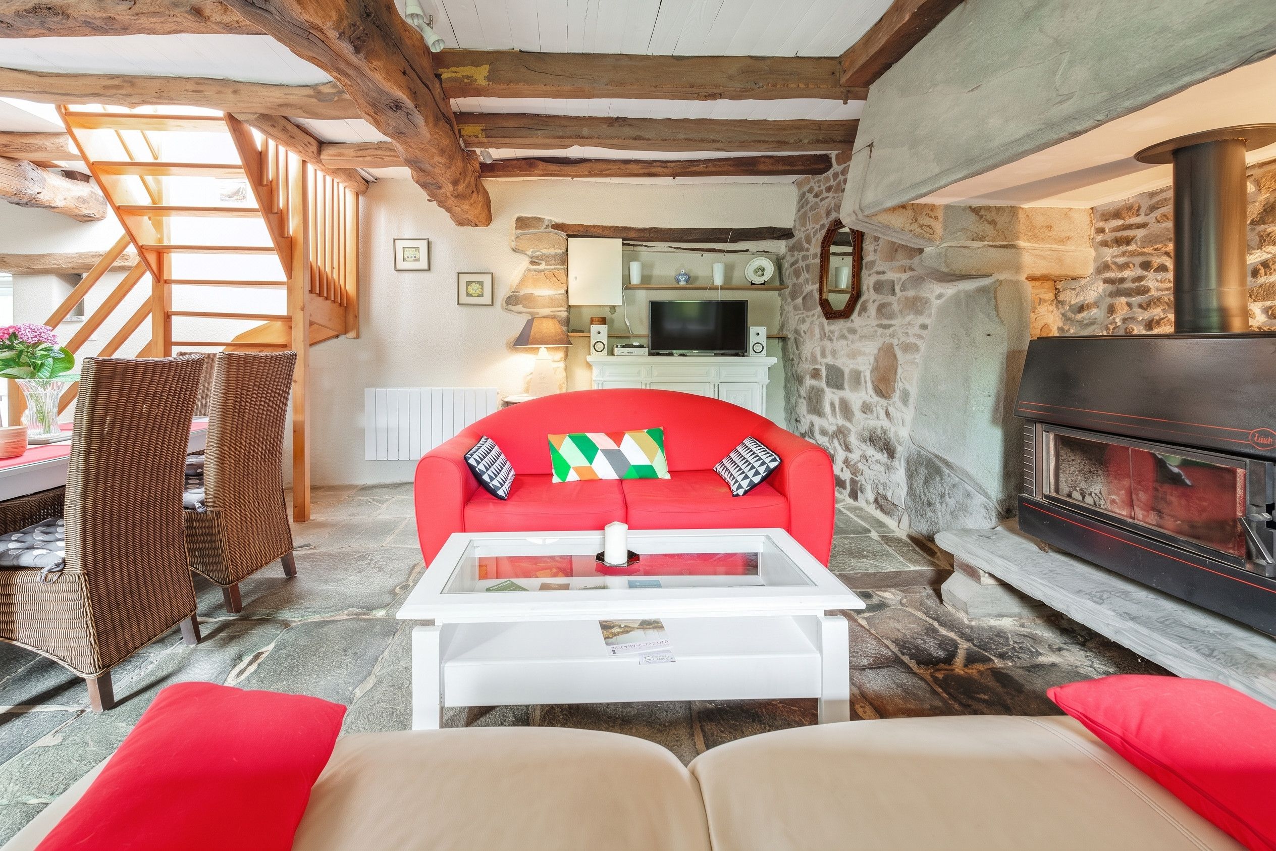 A cozy living room with a red sofa, a white coffee table, wicker chairs, a stone fireplace, and exposed wooden beams.