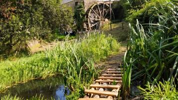 A rustic stone building with a large water wheel, surrounded by lush greenery and a wooden footbridge over a small stream.