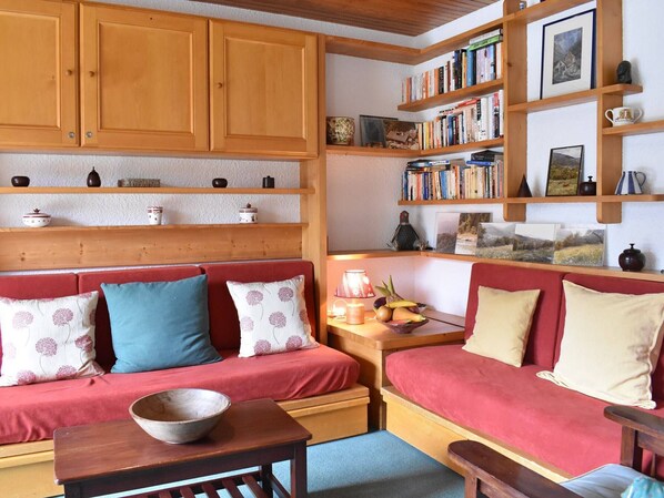 Un salon chaleureux avec un canapé rouge, des étagères en bois remplies de livres et d’objets décoratifs, et une petite table sur laquelle se trouve un bol.