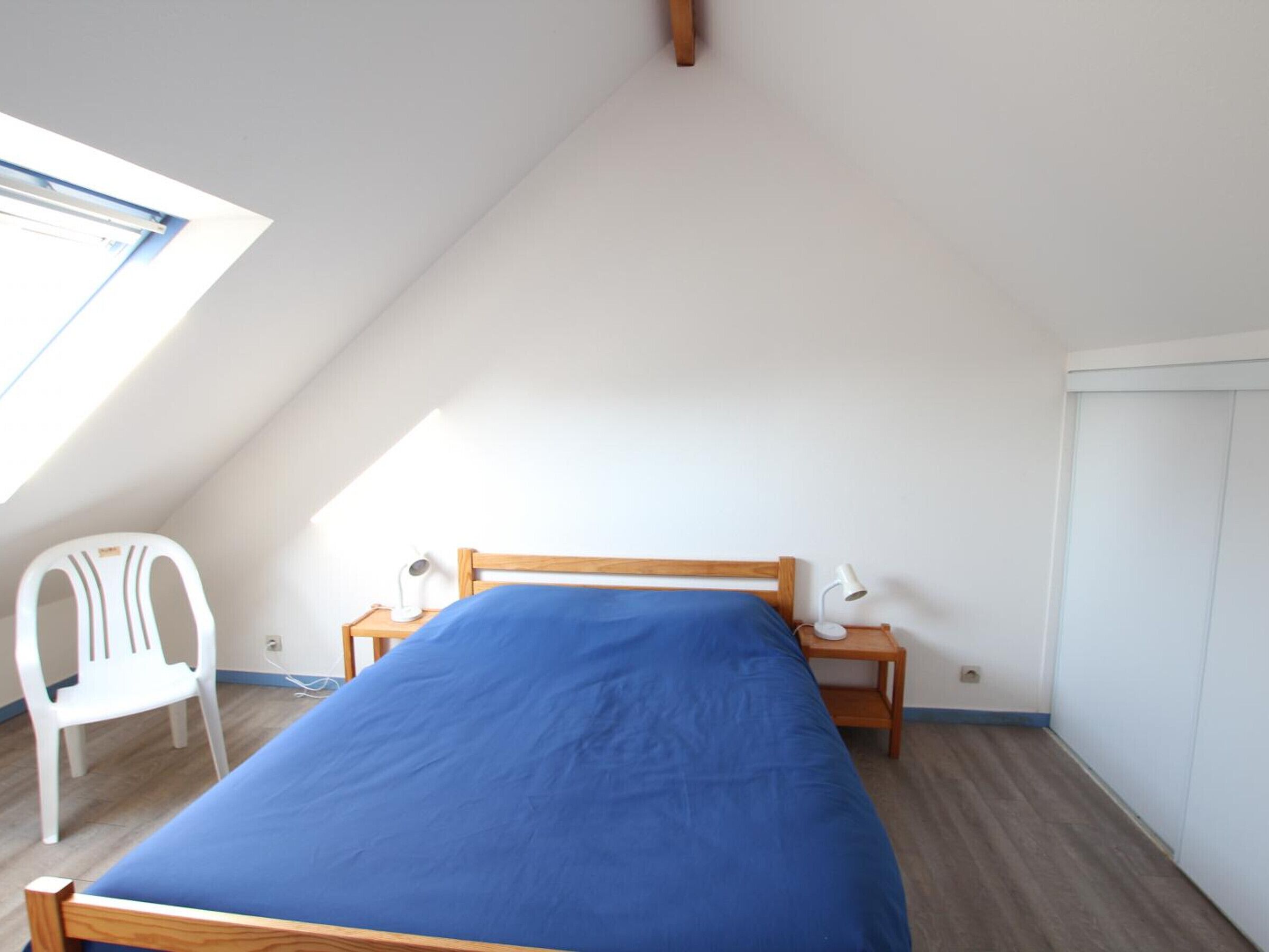 A small, single-bed room with a sloped ceiling, a wooden bed frame, a blue mattress, a white chair, a bedside table with a lamp, and a window providing natural light.
