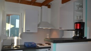 A compact kitchen with white cabinets, a built-in oven, a microwave, a coffee maker, and a window with blinds. - Charming Beachside House in Bréhal with Terrace, BBQ, and Wi-Fi (Bréhal-Saint-Martin de Bréhal)