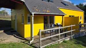 A yellow building with a covered porch and a wooden deck. - Charming Beachside House in Bréhal with Terrace, BBQ, and Wi-Fi (Bréhal-Saint-Martin de Bréhal)