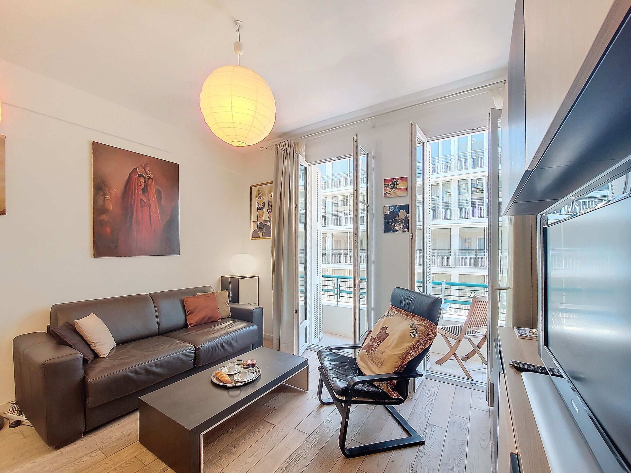 Ein modernes Wohnzimmer mit einem Ledersofa, einem Couchtisch mit einer Auswahl an Gebäck, einem schwarzen Sessel und einem großen Fenster mit Blick auf Gebäude.