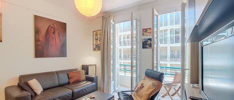 Ein modernes Wohnzimmer mit einem Ledersofa, einem Couchtisch mit einer Auswahl an Gebäck, einem schwarzen Sessel und einem großen Fenster mit Blick auf Gebäude.