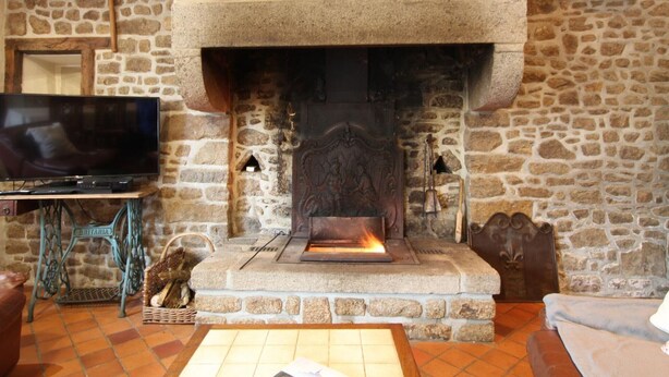 A stone fireplace with a television on a stand to the left, a basket of wood, and a table in front. - Spacious Cottage in South Manche with Fireplace and Private Garden (Le Grand-Celland)