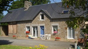 A stone building with a sloped roof, two doors, and a small window. There are outdoor chairs and a table in front. - Spacious Cottage in South Manche with Fireplace and Private Garden (Le Grand-Celland)