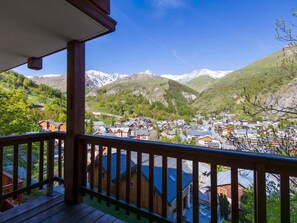 A wooden balcony overlooking a village with houses and snow-capped mountains in the background.