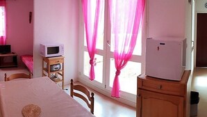 Une chambre à coucher avec un lit, une commode en bois, une petite télévision posée sur un meuble en bois, et une fenêtre avec des rideaux roses.