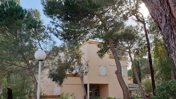 Un bâtiment à deux étages, dot é d’une façade beige et entouré de verdure et d’arbres.